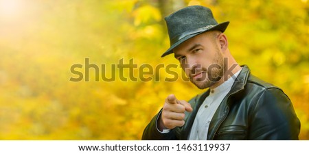 Man and autumn background. A brutal man with a beard, a black leather jacket and a hat shows a finger gesture forward. Banner