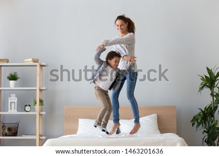 Happy african family mixed race ethnicity kid boy and mom baby sitter hold hands jumping on bed mattress, cheerful mother having fun laughing playing funny active game with cute child son in bedroom Royalty-Free Stock Photo #1463201636