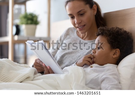 Mixed race african american family mom baby sitter and cute little kid son read book fairy tale in bed, cute small adopted child boy listening to mom foster parent learn reading in bedroom in morning Royalty-Free Stock Photo #1463201651