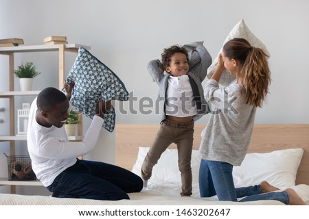 Happy mixed race ethnicity family playing pillow fight on bed, carefree african american parents having fun with cute little child kid son laughing in bedroom in the morning enjoy leisure activity