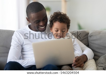 Cute small kid son learning using laptop watching cartoons with happy black dad, african american mixed race family father teaching child having fun with computer device sit together on couch at home