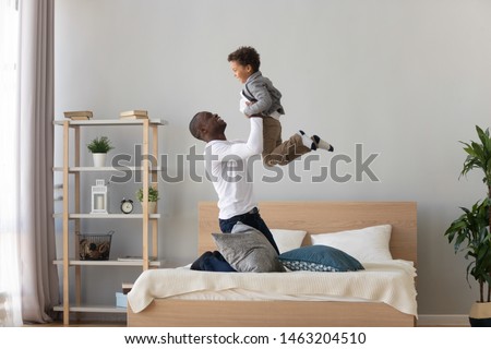 Happy family loving black father holding lifting cute small mixed race child son playing on bed, funny little kid boy flying in single african american dad arms having fun feel joy bonding in bedroom