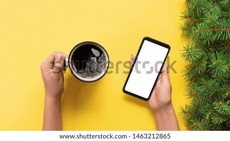 Female hands holding black mobile phone with blank white screen and mug of coffee. Mockup image with copy space. Top view banner on yellow background, flat lay.