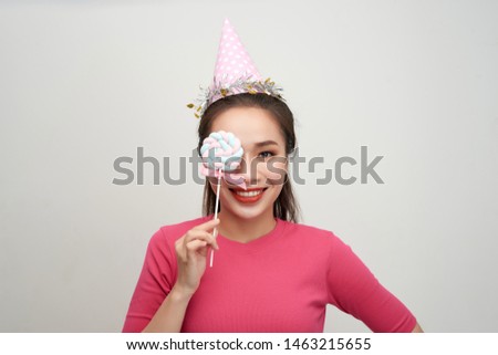 Portrait happy smiling woman in a birthday cap closes her eye with a lollipop on stick over pink background