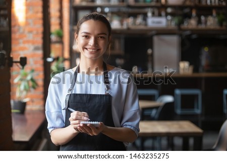Portrait of smiling millennial waitress standing wearing uniform holding notebook looking at camera, positive happy cafe or restaurant female staff in apron ready to take order. Good service concept Royalty-Free Stock Photo #1463235275