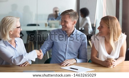 Smiling middle-aged female realtor handshake excited husband closing deal with overjoyed first time buyers couple, happy spouse shake hand signing agreement with banker take property loan or mortgage
