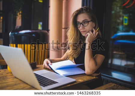 Serious woman in glasses with pensive look skilled copywriter reading article on laptop computer, sitting in coffee shop outdoors. Female marketing writer using notebook for searching information 