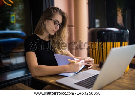 Female in glasses professional business content writer reading information from textbook during distance work on laptop computer while sitting in coffee shop during resting time in summer. 