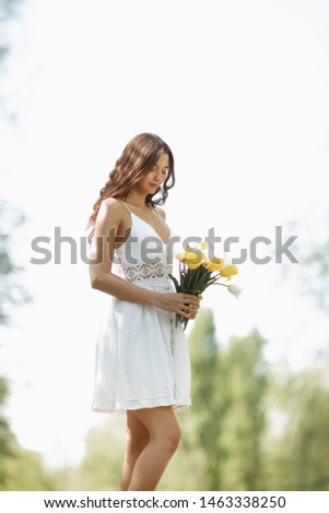 Attractive woman in white dress with a bouquet of spring flowers standing against nature bokeh green background. Creative photo of gorgeous young mixed race Caucasian Asian brunette girl holding