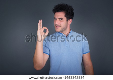 Glad attractive man shows ok sign with hand as expresses approval, has cheerful expression. Photo of handsome male has appealing appearance, being optimistic. Standing against gray wall.