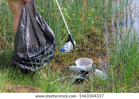 A Litter Picker Being Used To Pick Up Plastic Waste Washed Up Between The Reeds Of A lake.