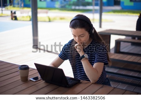 Pensive girl student, brunette, in headphones with a smartphone, a laptop and a paper cup of coffee sits at a wooden table. Writes a term paper. Front view. Royalty-Free Stock Photo #1463382506
