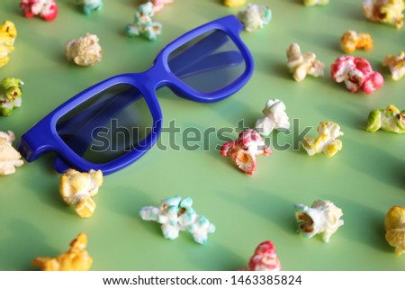 Glasses in blue plastic frame to watch the movie in 3D format among colored popcorn and candy on a green background.