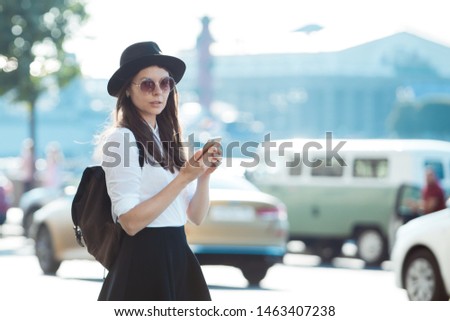 young woman in a hat and with a backpack walks in the city and uses a smartphone. Hipster on a walk uses the phone and takes photos for social networks