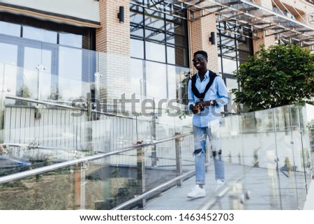 Portrait of young American man in blue shirt holding coffee while walking in the street. Model. Outfit.