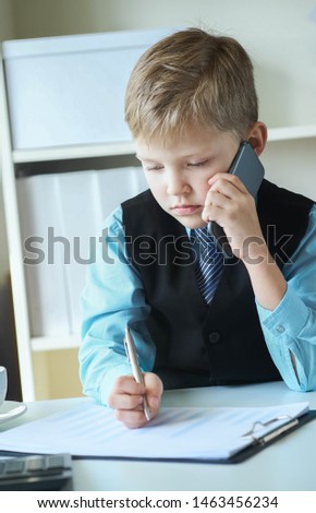 Little executive businessman boss boy making notes and talks on the phone and making notes sitting at the desk with laptop in office. Concept of early financial education