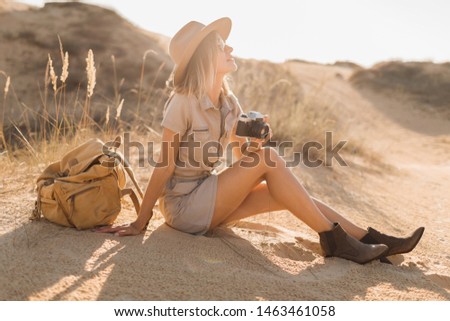 beautiful stylish young woman in khaki dress in desert traveling in Africa on safari wearing hat and backpack taking photo on vintage camera, exploring nature, sunny summer, traveler on vacation