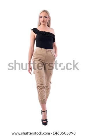 Elegant feminine blonde woman in beige trousers walking and looking at camera. Full body isolated on white background. 