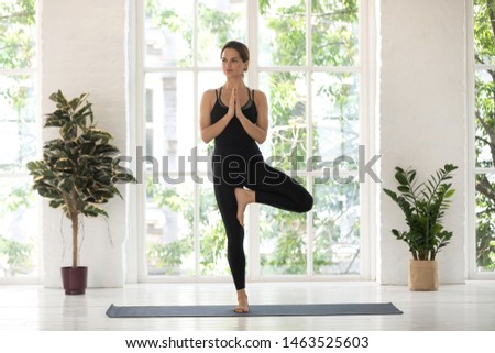 Woman wear black activewear practise yoga performs Tree pose standing on mat inside of cozy room with plants and greenery behind panoramic window Vrksasana Vriksasana asana, healthy lifestyle concept Royalty-Free Stock Photo #1463525603