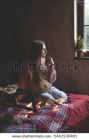 Caucasian young woman with long straight hair in a knitted sweater on the bed looks out the window and holds a mug with a drink. Christmas interior decor.