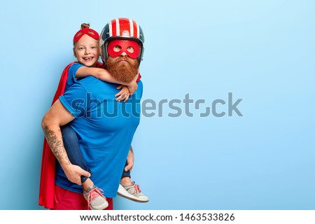 Horizontal shot of caring father gives piggyback to small child, play interesting superhero game, wear special costumes, pose against blue background with free space, have great impact on world Royalty-Free Stock Photo #1463533826