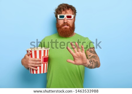 Displeased unhappy ginger guy in cinema refuses talk about film and characters after watching, makes rejection gesture, wears stereo glasses and holds bucket of junk food, isolated on blue background