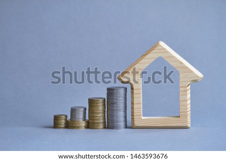 Wooden figurine of house with several columns of coins nearby on gray background, the concept of buying or renting a building, selective focus
