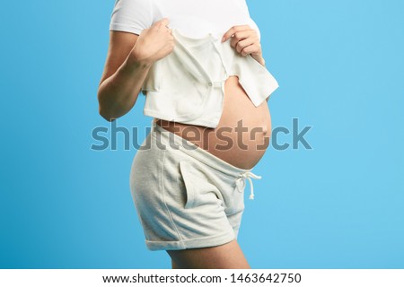 close up cropped photo. pregnant woman holding baby's clothes on her big belly. isolated blue background, studio shot.heir, successor