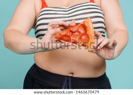 Overweight woman holding a pizza slice, midsection shot. Junk food, excess calories and health issues.