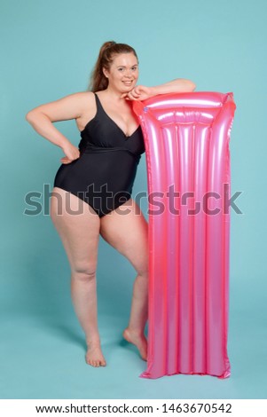 Happy overweight woman in a black swimsuit with a pink mattress. Overcoming body-insecurity of being fat. Full length portrait on blue background.