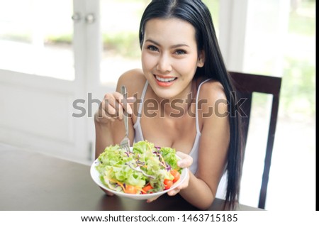 Young good health beautiful smiling Asian long hair girl is eating healthy food such as green salad sitting at home. She is looking at cabbage on her fork. Healthy food and diet concept.
