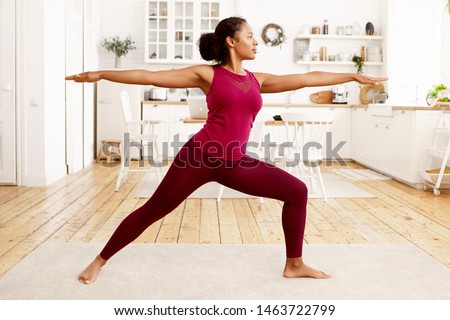 Fitness, training, health and wellness concept. Side view of athletic young Afro American housewife in stylish sportswear practicing yoga in the morning, doing warrior 2 pose on mat in kitchen Royalty-Free Stock Photo #1463722799