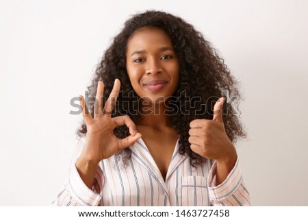 Joy, positiveness and body language. Beautiful happy young girl with curly black hair posing isolated dressed in silk pajamas, making thumbs up gesture and showing ok sign, smiling