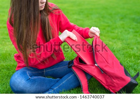 Cropped closeup photo of beautiful charming teen girl packing stuff into opened schoolbag sitting on green grass lawn inside the building outside