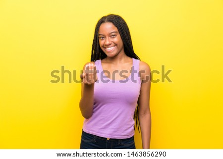 African American teenager girl with long braided hair over isolated yellow wall inviting to come with hand. Happy that you came