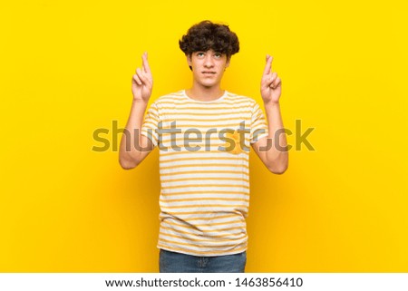 Young man over isolated yellow wall with fingers crossing and wishing the best