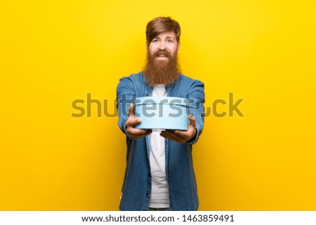 Redhead man with long beard over isolated yellow wall holding gift box