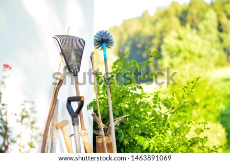 Old vintage gardening tools -  spade, fork and rake standing by white wall on the terrace with beautiful green garden in background. Rural countryhouse farm garden in gold sunset light. 
 Royalty-Free Stock Photo #1463891069
