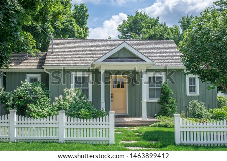 A small vintage cottage style home, in pastel green and yellow colours with white trim, set behind a white picket fence, surrounded by large mature trees, in a residential neighborhood.  Royalty-Free Stock Photo #1463899412