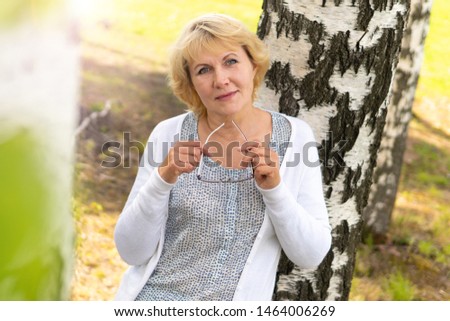 Middle-aged woman in the garden in nature. She stands by a birch tree and holds her glasses in her hands. The woman thought. She's looking at the camera.