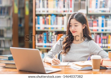 Asian young Student in casual suit doing homework and using technology laptop in library of university or colleage with various book and stationary over the book shelf background, Back to school Royalty-Free Stock Photo #1464037382