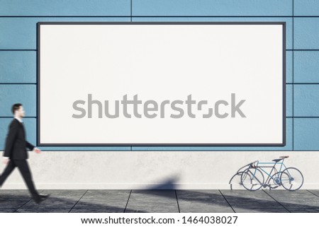 Businessman walking past empty blue exterior wall with bicycle on street, empty poster and sunlight. Mock  up,