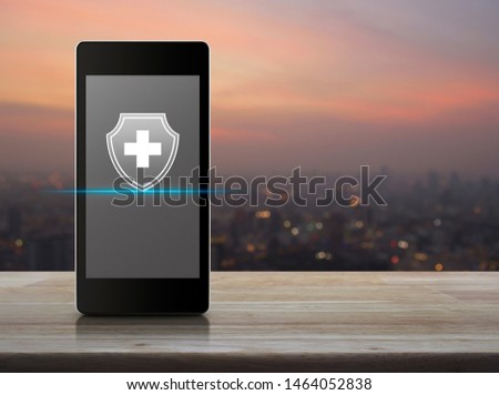 Cross shape with shield flat icon on modern smart mobile phone screen on wooden table over blur of cityscape on warm light sundown, Business healthy and medical care insurance online concept