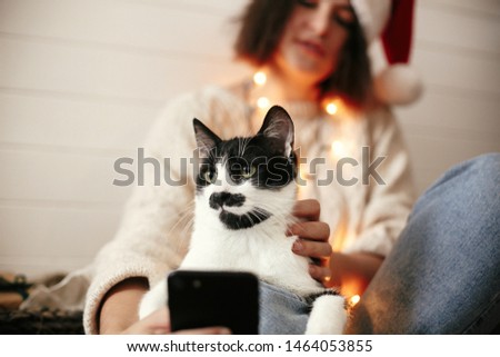 Stylish happy girl in santa hat taking selfie with cute cat in christmas lights on background of gifts. Young hipster woman in sweater hugging kitty with funny emotions at christmas tree