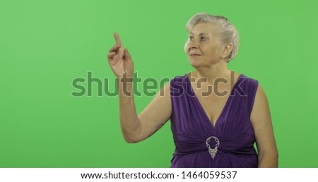 An elderly woman points at something and smiles. Old pretty grandmother in a purple dress. Place for your logo or text. 