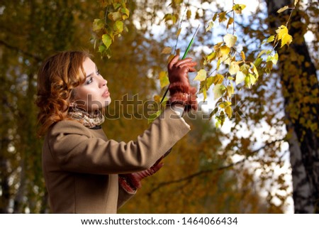 Young beautiful woman with palette and brush paints colors leaves on the trees. Outdoors in autumn park.