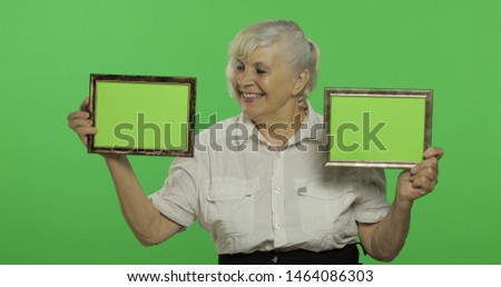 An elderly woman holding two frames with green image. Old pretty grandmother in a white shirt. Place for your logo or text. 