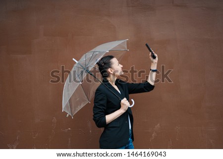 A young attractive woman in a black jacket and blue jeans posing outdoor against the background of the building. Holds a transparent umbrella. Rainy weather.