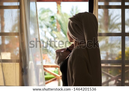
Young woman, wearing white bathrobe and hair towel, standing by the panoramic window with tulle curtains watching beautiful palms resort view. Silhouette of pretty girl at the tropical hotel balcony.