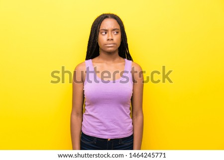 African American teenager girl with long braided hair over isolated yellow wall making doubts gesture looking side